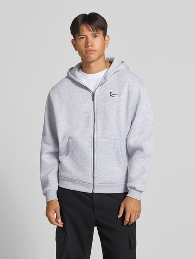 Männermodel trägt Sweatjacke mit Label-Stitching und Känguru-Tasche von Karl Kani, hellgrau, Regular Fit, Baumwoll-Mix, Ganzkörperansicht von vorne.