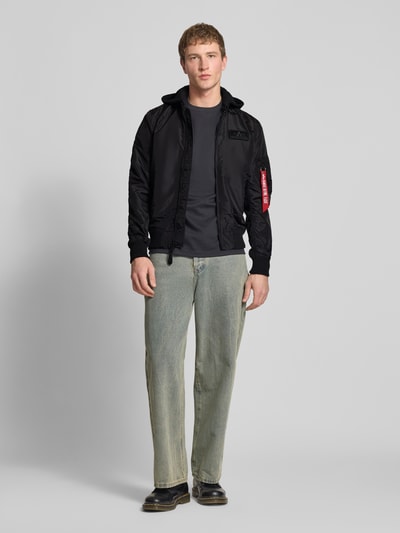 Männermodel trägt schwarzen Blouson mit Logo-Anhänger Modell 'MA-1 TT' von Alpha Industries, lockere Passform, Kapuze aus Sweat, Reißverschluss, Armtasche, Label-Patch, elastische Abschlüsse, Rückenlänge bei Größe M 69 cm, Schulterbreite 46 cm, Brustweite 54 cm, Ärmellänge 67 cm, Ganzkörperansicht von vorne