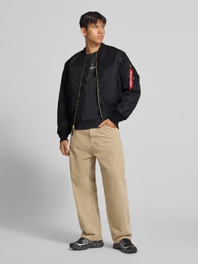 Männermodel trägt Bomberjacke mit Pattentaschen und Label-Applikation von Alpha Industries in Schwarz, Reißverschluss, Ganzkörperansicht von vorne. Regular Fit, Stehkragen, Rückenlänge 69 cm, Schulterbreite 44 cm, Brustweite 54 cm, Ärmellänge 64 cm.