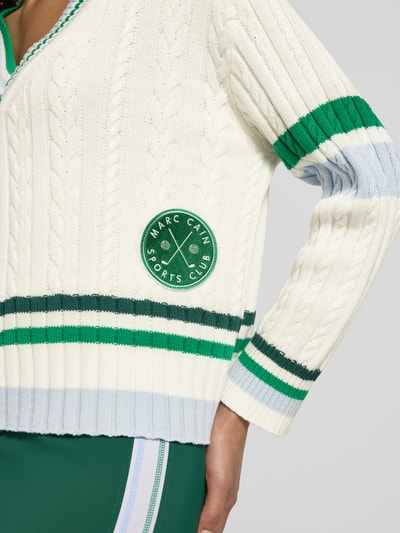 Frauenmodel trägt Relaxed Fit Pullover mit Lanawoll-Anteil von Marc Cain, cremefarben mit grünen und blauen Streifen, Nahaufnahme Oberkörper. Baumwoll-Mix, V-Ausschnitt, Logo-Stitching, sportliches Design.