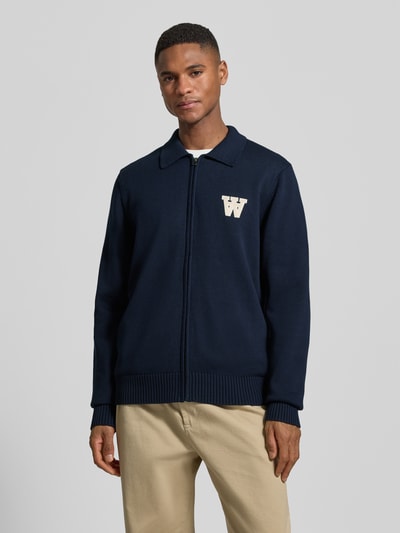 Männermodel trägt Regular Fit Cardigan in Strick-Optik Modell 'Tay' von Wood Wood, marineblau, Logo-Stitching, Reißverschluss, Ganzkörperansicht von vorne.