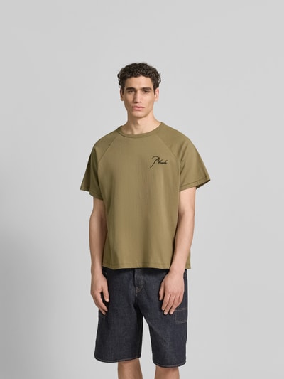 Männermodel trägt Regular Fit T-Shirt aus reiner Baumwolle von RHUDE in Oliv mit Label-Stitching, Raglanärmel, Rundhalsausschnitt, Ganzkörperansicht von vorne.