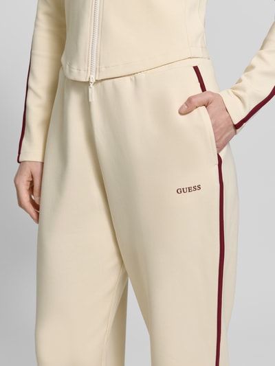 Kobiece modelka nosi Jogpants mit Logo Modell 'JASMINE' od Guess Activewear, kremowy kolor, materiał z domieszką lyocellu, krój regular fit, kieszenie, logo, zdjęcie: zbliżenie na górną część nogawek z przodu.