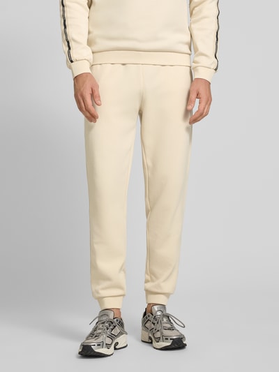 Männermodel trägt Sweatpants aus Baumwoll-Mix von EA7 Emporio Armani in Beige mit elastischem Bund, Eingrifftaschen, lockere Passform, Ganzkörperansicht von vorne.