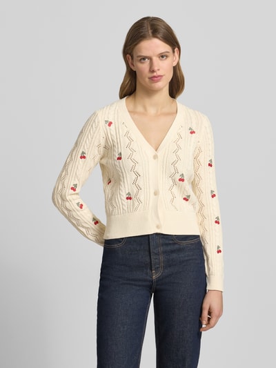 Modelka kobieca nosi Regular Fit Strickjacke aus Baumwoll-Mix Modell 'BIRTIE' od Only, jednokolorowy kremowy kardigan rozpinany ze ściegiem warkoczowym, kwiatowymi haftami, gładka dzianina, zapięcie na listwę guzikową, krój regular fit, głęboki dekolt w serek, długi rękaw, prążkowane wykończenie, mieszanka bawełny, faktura dzianiny, widok całej sylwetki z przodu.