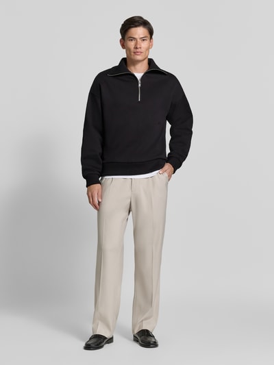Männermodel trägt Regular Fit Sweatshirt aus Baumwoll-Mix mit Troyer-Kragen von DRYKORN, schwarze Farbe, gerade Formen, glatte Textur, Logo-Stitching, kombiniert mit heller Stoffhose und schwarzen Schuhen, Ganzkörperansicht von vorne. Rückenlänge 64 cm, Ärmellänge 57 cm.
