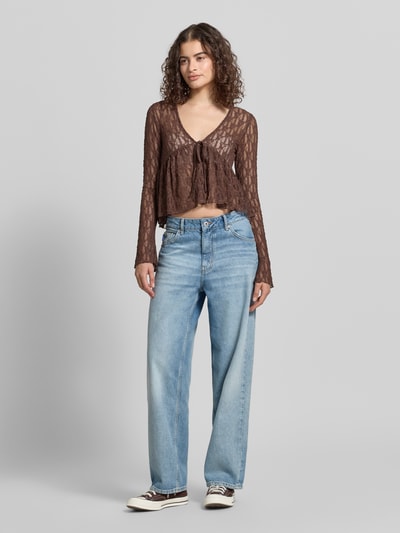 Frauenmodel trägt Shaped Fit Blusentop mit Bindeverschluss Modell 'LAYA' von Only, Semitransparent Optik, Mesh, abstraktes Muster, langarm, tiefer V-Ausschnitt, ohne Kragen, Taillengürtel, Materialverarbeitung: Mesh, braune Töne, lockere Passform, Jeans kombiniert, Ganzkörperansicht von vorne.