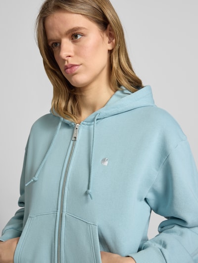 Kobieta nosi bluzę rozpinaną z kapturem z bawełny, krój loose fit, model 'Casey', wyhaftowane logo Carhartt Work In Progress, wpuszczane kieszenie, elastyczne wykończenia, jasnoniebieski kolor, zamek błyskawiczny, damska bluza rozpinana z wyhaftowanym logo model 'Casey' od Carhartt Work In Progress, zbliżenie na górną część ciała