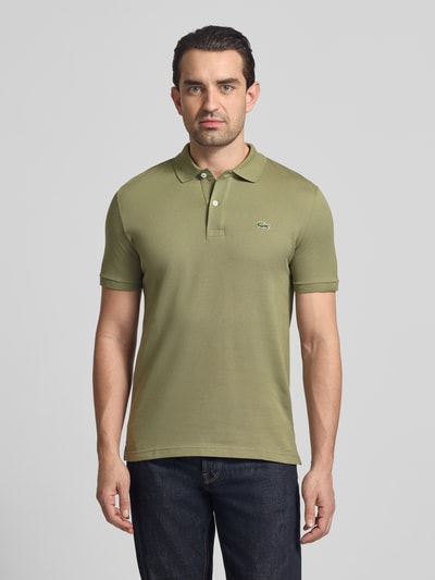 Mężczyzna nosi Slim Fit Poloshirt aus reiner Baumwolle von Lacoste, jednokolorowy, krój slim fit, wyhaftowane logo, kołnierzyk polo, krótki rękaw, prążkowane zakończenia, wykończenie z rozcięciami bocznymi, pika, bawełna. Ganzkörperansicht von vorne.