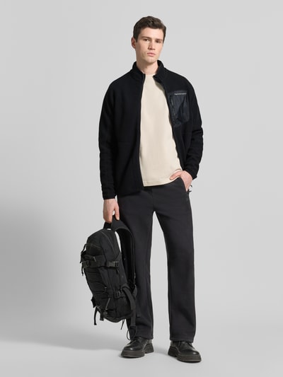 Mężczyzna nosi Fleecejacke mit Zweiwege-Reißverschluss Modell 'NatureRush' od PEAK PERFORMANCE, czarny kolor, krój regular fit, stójka, kieszeń na piersi, widoczna faktura materiału, widok całej sylwetki z przodu.