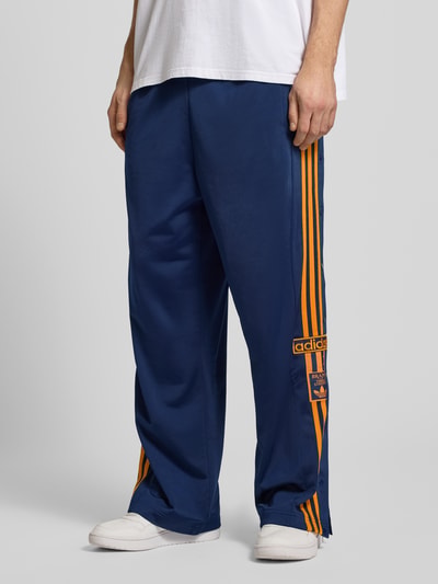 Männermodel trägt Sweatpants mit elastischem Bund von adidas Originals in Dunkelblau mit orangenen Streifen, Lockerer-Schnitt, Eingrifftaschen, Label-Stitching, Innenbeinlänge bei Größe M: 75 cm, Ganzkörperansicht von vorne.