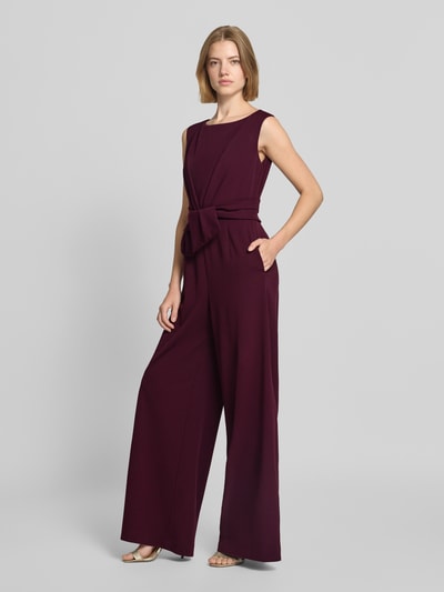 Frauenmodel trägt Regular Fit Jumpsuit in Wickel-Optik von Vera Mont in Dunkellila mit Bindegürtel und weitem Bein, Ganzkörperansicht von vorne.