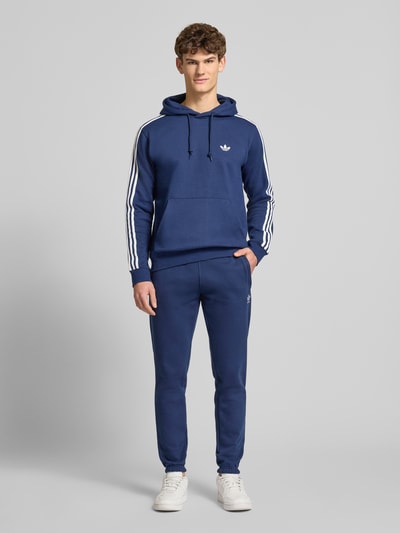 Model męski nosi Hoodie mit Kapuze od adidas Originals, bawełniany miks, krój luźny, kaptur, tunel, haftowane logo, kieszeń kangurka, ściągacze, długość tyłu w rozmiarze M 70 cm, długość rękawa w rozmiarze M 67 cm, widok całej sylwetki z przodu.