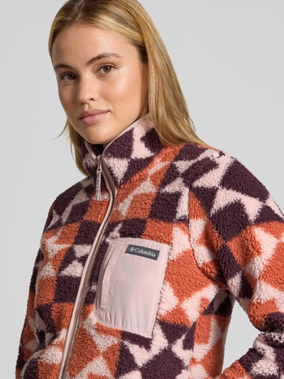 Modelka nosi Regular Fit Jacke mit Stehkragen od Columbia w geometryczne wzory, różowe i fioletowe kolory, kieszeń na piersi, krój regular fit, zamek błyskawiczny, polar, długość tyłu w rozmiarze S: 61 cm, szerokość ramion w rozmiarze S: 39 cm, szerokość klatki piersiowej w rozmiarze S: 49 cm, długość rękawa w rozmiarze S: 63 cm, zbliżenie na górną część ciała.
