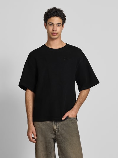 Männermodel trägt Relaxed Fit T-Shirt mit Strukturmuster von KARL KANI, Reine Baumwolle, Logo-Stitching, Rundhalsausschnitt, Ganzkörperansicht von vorne. Rückenlänge bei Größe M: 71 cm, schwarze Farbe, schlichte Struktur.
