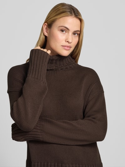 Frauenmodel trägt Relaxed Fit Strickpullover aus reiner Baumwolle mit Rollkragen von MOS MOSH, dunkles Braun, grob gestrickt, Nahaufnahme Oberkörper. Rücken- und Ärmellänge bei Größe S 62 cm, elastische Abschlüsse, entspannte Passform.