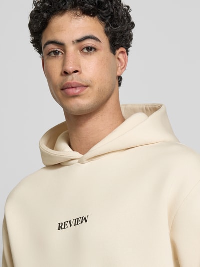 Männermodel trägt Regular Fit Hoodie mit Logo-Stitching von REVIEW in Beige, glatte Struktur, florales Stitching, Nahaufnahme Oberkörper.