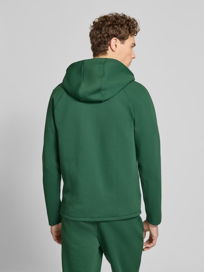 Męski model nosi Regular Fit Sweatjacke mit Baumwoll-Anteil od Lacoste Sport w jednolitym zielonym kolorze, krój regular fit, materiał bawełniany, kaptur ze sznurkiem, długi rękaw, kieszenie boczne, zdobiona naszywką z logo, widok całej sylwetki z tyłu.