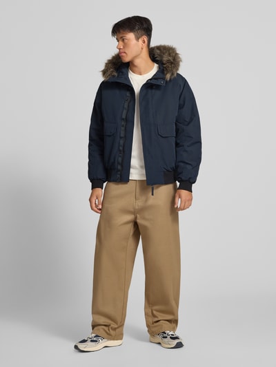Männermodel trägt Bomberjacke mit Label-Patch Modell 'Everest' von Superdry in Dunkelblau mit Kunstfellkapuze, Ganzkörperansicht von vorne. Regular Fit, Reiß- und Druckknopfverschluss, Pattentaschen, neutrale Farbtöne, glatte und flauschige Texturen, Rückenlänge 71 cm, Schulterbreite 50 cm, Brustweite 65 cm, Ärmellänge 70 cm.