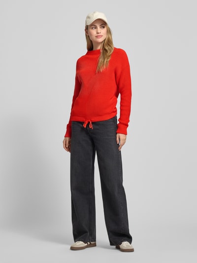 Kobiece modelka nosi Strickpullover mit Stehkragen und Tunnelzug od Jake*s Casual, czysty bawełna, krój regular fit, długość tyłu S: 67 cm, długość rękawa S: 56 cm, czerwony, faktura prążkowana, luźne czarne spodnie, białe sneakersy, pełny widok od przodu.