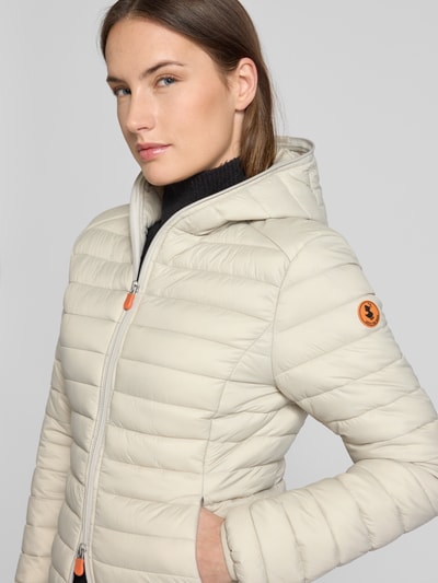 Frauenmodel trägt Steppjacke mit Kapuze Modell 'DAISY' von SAVE THE DUCK in Beige mit orangem Logo, körpernahe Passform, Nahaufnahme Oberkörper