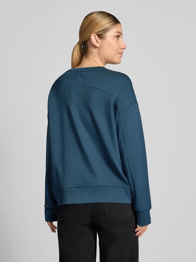 Modelka nosi bluzę 'Sweatshirt mit Rundhalsausschnitt und Label-Detail' od SCHIESSER, mieszanka modalu z dodatkiem streczu, krój loose fit, okrągły dekolt, opadające ramiona, detal z logo, matowa gładka tekstura w kolorze niebieskim, widok całej sylwetki od tyłu.