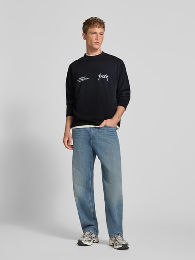 Männermodel trägt Regular Fit Sweatshirt aus Baumwoll-Mix Modell 'SRC® Pacer' von ANOTHER COTTON LAB, schwarz mit Statement-Print, Strukturmuster, Stehkragen, kombiniert mit blauer Jeans, Ganzkörperansicht von vorne.