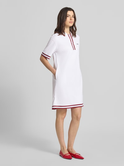 Frauenmodel trägt knielanges Polokleid mit Label-Stitching Modell 'Tasa' von JOOP!, weiß mit roten Kontraststreifen, Baumwoll-Elasthan-Mix, Gerader-Schnitt, Ganzkörperansicht von vorne.