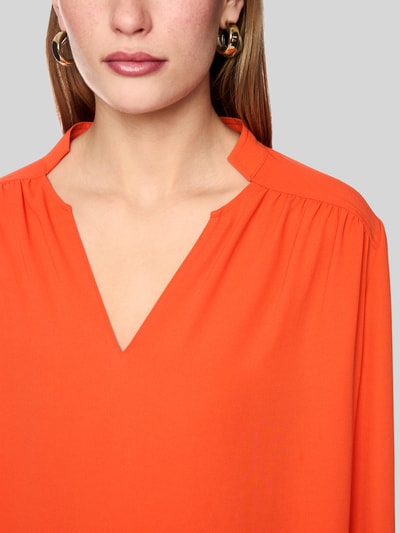 Vrouwenmodel draagt Regular Fit Bluse mit V-Ausschnitt van Marc Cain in effen oranje met opstaande kraag en plooien, Nahaufnahme Oberkörper. Ruglengte maat 36: 63 cm, mouwlengte maat 36: 65 cm.