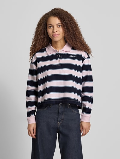 Frauenmodel trägt Cropped Poloshirt mit Label-Stitching von Guess Jeans in Dunkelblau, Rosa und Weiß, Reine Baumwolle, Knopfleiste, Longsleeve, Ganzkörperansicht von vorne.
