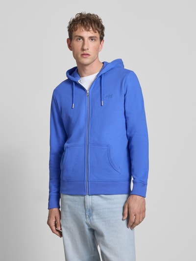 Männermodel trägt Sweatjacke mit Kapuze und Logo-Stitching von Superdry aus Baumwoll-Mix, Regular-Fit in Blau mit Eingrifftaschen, Ganzkörperansicht von vorne.