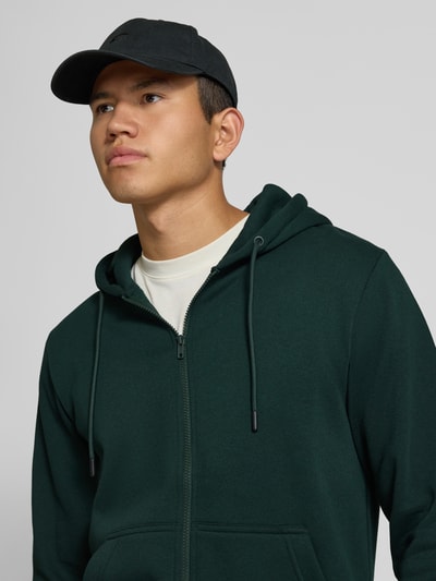 Männermodel trägt Regular Fit Sweatjacke aus Baumwoll-Mix Modell 'BRADLEY' von JACK & JONES in Dunkelgrün, Nahaufnahme Oberkörper. Baumwoll-Mix, Kapuze mit Tunnelzug, getrennte Känguru-Tasche, elastische Abschlüsse.