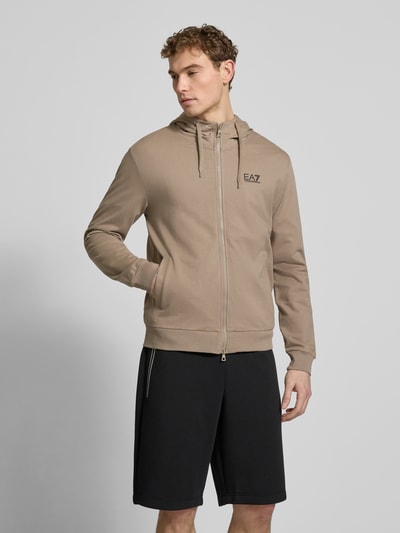 Męski model nosi Slim Fit Sweatjacke mit Kapuze od EA7 Emporio Armani, beżowy, dresowy materiał, logo, kieszenie, widok całej sylwetki z przodu.
