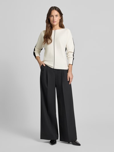 Frauenmodel trägt Regular Fit Pullover aus Viskose-Mix Modell 'Dezember' von More & More in Strick-Optik, cremefarben mit schwarzen Akzenten, kombiniert mit weiter schwarzer Hose, U-Boot-Ausschnitt, Ganzkörperansicht von vorne.