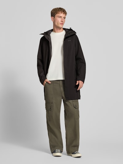 Männermodel trägt Regular Fit Mantel mit Kapuze Modell 'Brussel' von Helly Hansen in Schwarz, Logo-Detail, Ganzkörperansicht von vorne. Rückenlänge bei Größe M: 96 cm, Schulterbreite: 46 cm, Brustweite: 51 cm, Ärmellänge: 66 cm.