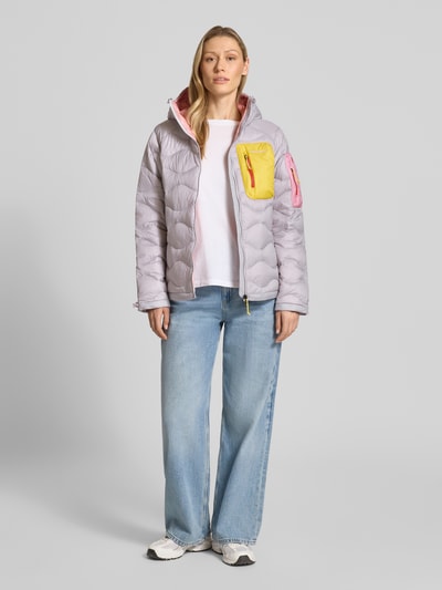 Frauenmodel trägt Jacke mit Entendaunen-Füllung und Kapuze von PEAK PERFORMANCE in Hellgrau mit gelber und rosa Tasche, prosty krój, kaptur z troczkami, zamek błyskawiczny, wyhaftowane logo, brustasza zapinana na zamek błyskawiczny, kieszeń zapinana na zamek błyskawiczny na rękawie, boczne kieszenie zapinane na zamek błyskawiczny, regulowane wykończenia z troczkami, długość tyłu w rozmiarze S: 65 cm, szerokość ramion w rozmiarze S: 40 cm, szerokość klatki piersiowej w rozmiarze S: 51 cm, długość rękawów w rozmiarze S: 63 cm, Ganzkörperansicht von vorne