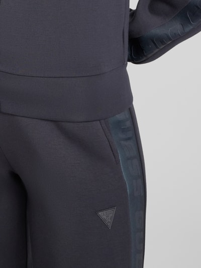 Kobiece modelka nosi Sweatpants mit elastischem Bund Modell 'ALLIE' od Guess Activewear, ciemnoszary kolor, luźny krój, francuskie kieszenie, wiskoza ze stretchem, szeroki pas i tunel na sznurek, widok z przodu, zbliżenie na pas i kieszeń.