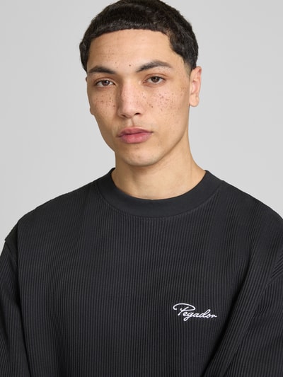 Męski model nosi Oversized T-Shirt mit Strukturmuster von Pegador, mieszanka bawełny, krój oversized fit, okrągły dekolt, wyhaftowane logo, długość tyłu w rozmiarze M: 71 cm, fakturowana powierzchnia, ciemny kolor, nahaufnahme górnej części ciała.
