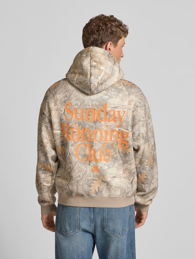 Männermodel trägt Hoodie mit Logo- und Motiv-Print von ANOTHER COTTON LAB in beige-braunen Tönen, Baumwoll-Mix, Loose Fit, Ganzkörperansicht Rückansicht. Rückenlänge 63 cm, Känguru-Tasche, Label-Patch, Kapuze, Ärmellänge 51 cm.