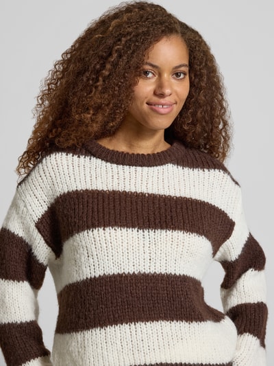 Modelka nosi sweter Relaxed Fit Pullover im Chunky Knit Look model 'DORTHEA' od PIECES, biało-brązowe grube paski, faktura dzianiny, zbliżenie na górę ciała.