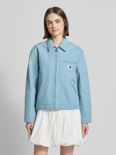 Frauenmodel trägt Jacke mit Label-Detail von Carhartt Work In Progress in Hellblau, Reine Baumwolle, Relaxed-Fit, Reißverschluss, Eingrifftaschen, Umlegekragen, Ganzkörperansicht von vorne. Rückenlänge bei Größe S: 58 cm, Schulterbreite: 47 cm, Brustweite: 54 cm, Ärmellänge: 60 cm.