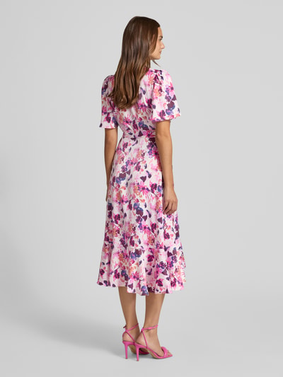 Frauenmodel trägt Wickelkleid in Midilänge Modell 'Thea' von YAS mit floraler Allover-Print, Zierschleife, Taillierte Passform, Elastischer-Material-Mix und Kurzärmelig, Farbtöne in Rosa und Violett, Ganzkörperansicht Rückansicht.
