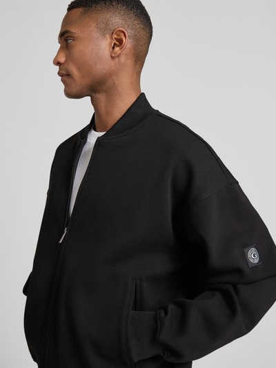 Männermodel trägt schwarze Sweatjacke mit Reißverschluss von Guess aus Baumwoll-Mix, Regular-Fit, gerippte Abschlüsse, Stehkragen, Eingrifftaschen, seitliche Nahaufnahme Oberkörper.