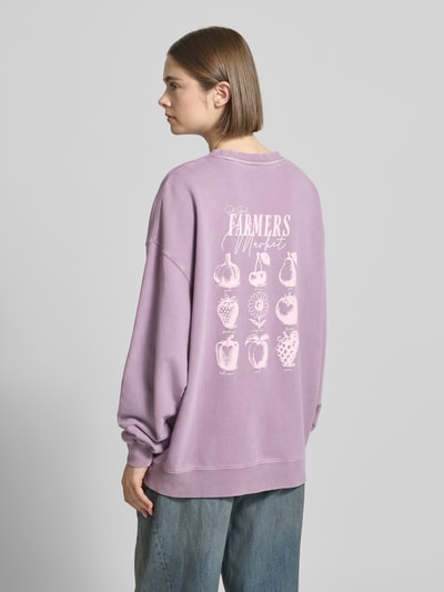 Frauenmodel trägt Oversized Sweatshirt mit Logo-Stitching Modell 'Farmers Market' von OH APRIL, Reine Baumwolle, Oversized-Fit, Gerippter Rundhalsausschnitt, überschnittene Schultern, Rückseitiger Statement-Print in Flieder mit weißen Prints, weiche Textur, Ganzkörperansicht Rückansicht.