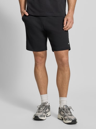 Männermodel trägt Sweatshorts mit Eingrifftaschen von Christian Berg Men, schwarze Farbe, Reine Baumwolle, Regular-Fit, Label-Detail, Nahaufnahme Oberschenkel bis Schuhe, Bundweite bei Größe M 41 cm, Innenbeinlänge 22 cm.