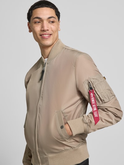 Męski model nosi Regular Fit Jacke mit Stehkragen od Alpha Industries, beżowy kolor, kieszeń na ramieniu, elastyczne ściągacze, zamek, widok z przodu, całkowity kontekst: długość pleców w rozmiarze M: 66 cm, szerokość ramion w rozmiarze M: 48 cm, szerokość klatki piersiowej w rozmiarze M: 54 cm, długość rękawa w rozmiarze M: 68 cm, krój regular fit
