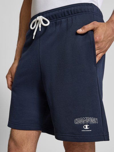 Männermodel trägt Sweatshorts mit Logo und elastischem Bund von CHAMPION, dunkelblauer Baumwoll-Mix, sichtbarer Tunnelzug, Nahaufnahme Oberkörper.