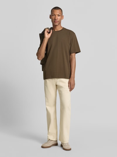 Männermodel trägt Oversized T-Shirt aus reiner Baumwolle von Lindbergh in Braun, halbärmelig, kombiniert mit heller Hose, Ganzkörperansicht von vorne, Kontext: Herren T-Shirt von Lindbergh, reine Baumwolle, Oversized, leicht überschnittene Schultern, halbärmeliges Design, Rückenlänge bei Größe M: 70 cm.
