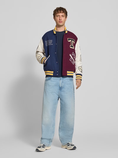 Männermodel trägt Regular Fit Bomberjacke aus Woll-Mix Modell 'LETTERMAN' von Tommy Jeans in Blau, Weiß und Bordeaux, Herren Bomberjacke, Ganzkörperansicht von vorne.