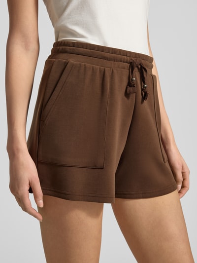 Frauenmodel trägt braune Sweatshorts mit Seitentaschen von Scotch & Soda, gerader Schnitt, elastischer Bund, Nahaufnahme Oberschenkel.